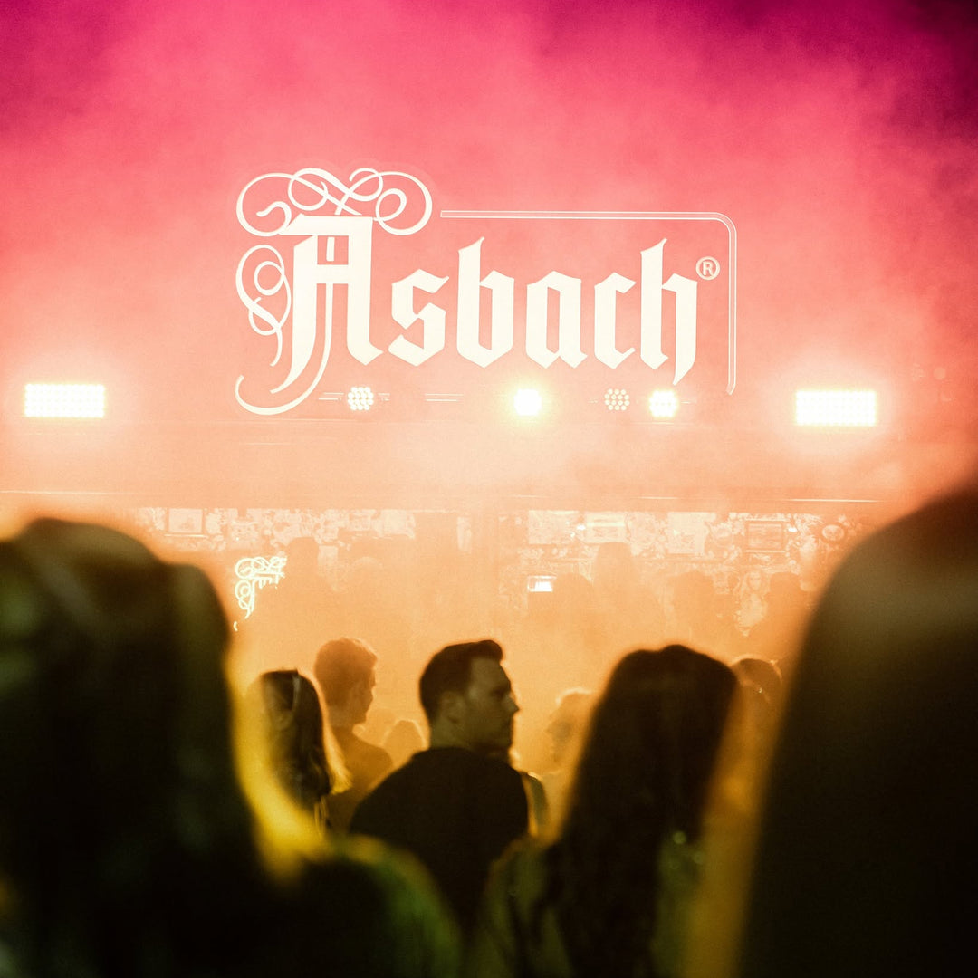 A concert or event scene with a large white 'Asbach' logo centered against pink and orange stage lighting, with silhouetted people in the foreground and smoke filling the background.