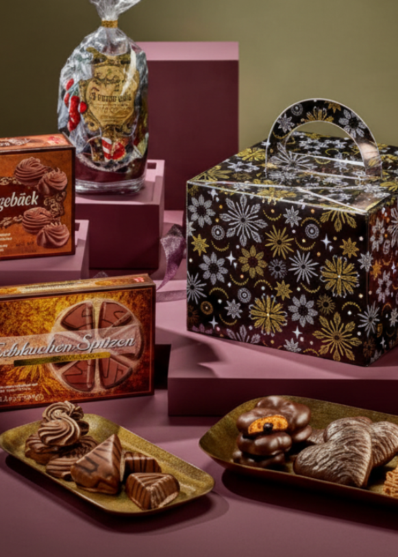 A promotional display of assorted packaged sweets and gift boxes arranged on a mauve-colored surface, with a product details panel on the right showing text and ingredients. The scene includes multiple branded boxes, a wrapped item, and plated pastries.
