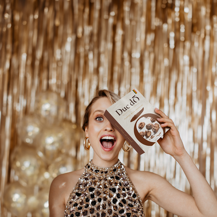 Woman holding a Duc d’O dark flaked truffles box against a gold party backdrop