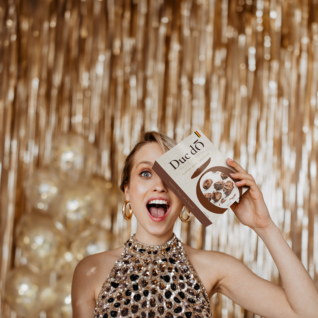 Woman holding a Duc d’O milk hazelnut flaked truffles box against a gold party backdrop