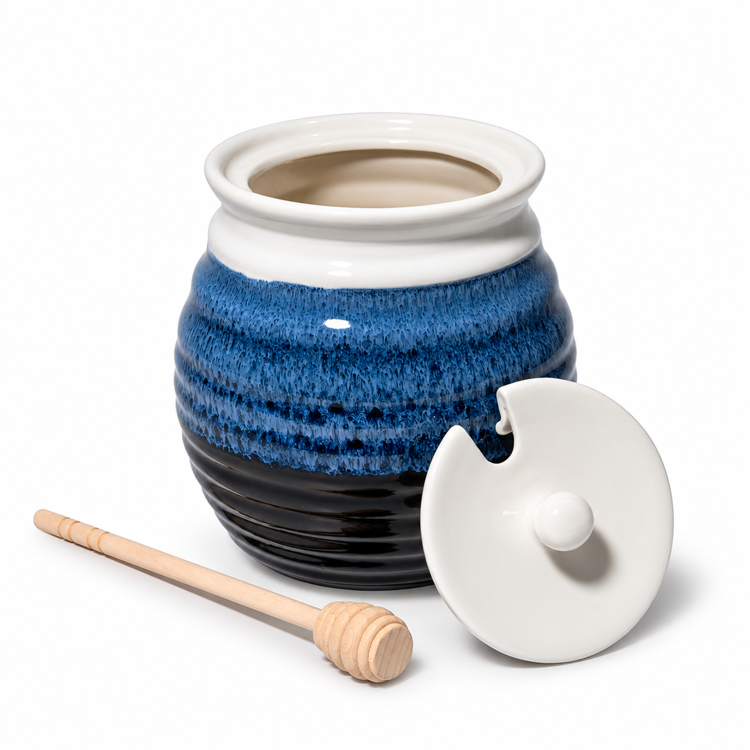 Blue glazed ceramic honey pot with white lid and wooden honey dipper on a white background